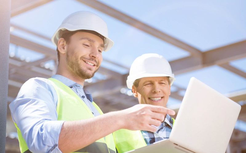 Two workers on a construction site with safety helmets on and a hi-vis vest both looking at a laptop
