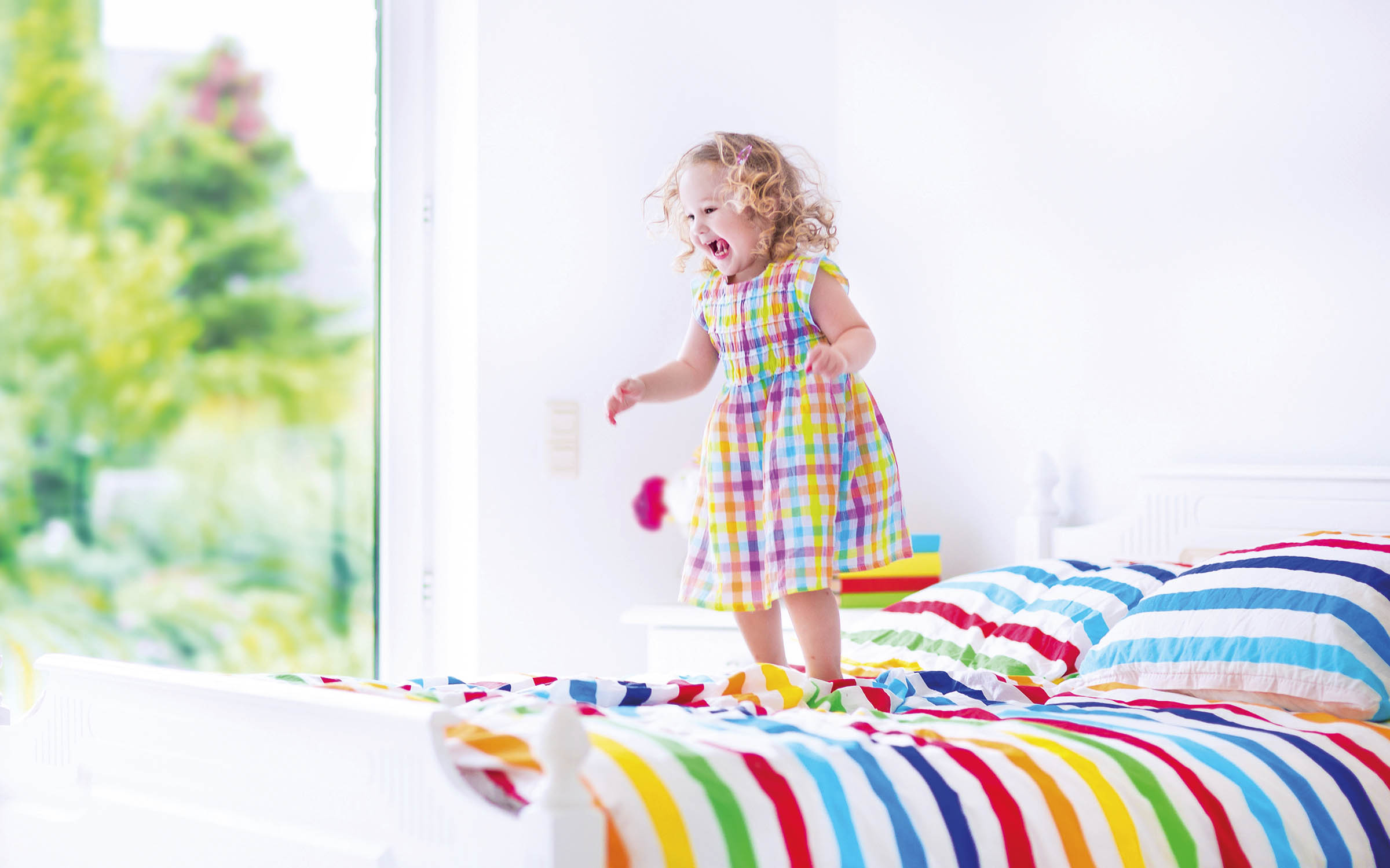 Child in coloured dress jumping on a colourful striped bed.